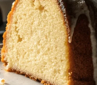 A sliced cream cheese pound cake with a golden crust and a drizzle of icing, displayed on a plate near a window.