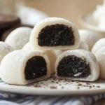 A white plate with several round Oreo mochi balls dusted with powdered sugar, some cut open to reveal a black Oreo filling, with whole Oreo cookies in the background.