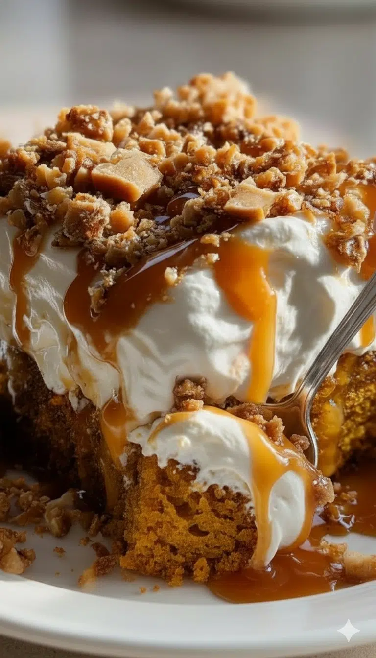 A close-up shot of a slice of pumpkin cake topped with whipped cream, caramel drizzle, and crunchy nut crumble, with a spoon taking a scoop.